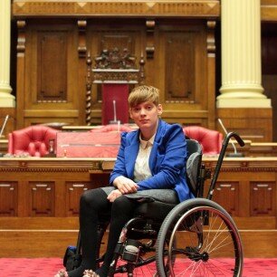 This is a photograph of Kelly Vincent in the Legislative Council. Ms Vincent is sitting in her manual wheelchair. She is wearing a white blouse and grey skirt with a blue blazer over the top and black stockings. The President's chair the Legislative Council can be seen in the background.