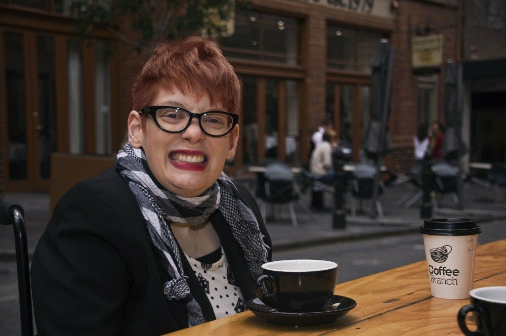 This is a photograph of Kelly Vincent enjoying a coffee on Leigh Street. In the photo she is smiling toward the camera and wearing a black jacket and grey scarf. She has short red hair, dark rimmed glasses, and bright red lipstick. In the background are some tables and chairs of a cafe behind her.