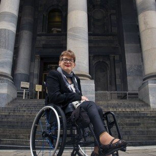 Kelly This is a photograph of Kelly Vincent in front of the steps of Parliament House. Ms Vincent is sitting in her manual wheelchair. She is wearing dark framed spectacles and red lipstick. She is wearing a white blouse and black pants. She also wears a black blazer and a black and white gingham print scarf. The steps of Parliament can be seen in the background.Vincent outside Parliament House