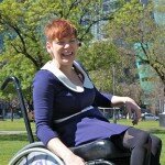 This is a picture of Ms Vincent in Victoria Square, Adelaide. Ms Vincent has short red hair, is wearing a blue dress and is using a manual wheelchair. She is on the lawns of VIctoria Square and the Federal Court building is in the background.