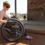 This is a photograph of Ms Vincent visiting the APY Lands. Ms VIncent is sitting in a three-wheeled, all-terrain, amnual wheelchair outside of the community shop in Kaltjiti. The front of the shop is decorated with a mural of indigenous art, depicting the local environment. There is a large step to access the entrance of the shop. There is a small balck dog in the background.