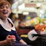 This is a picture of Ms Vincent at the Adelaide Central Market. Ms Vincent has short red hair, is wearing a blue dress, and is using a manual wheelchair. She is sitting next to a fruit stall stocked with red and green apples. She is holding a red apple and laughing.