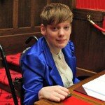 This is a picture of Ms Vincent in the South Australian Legislative Council. Ms Vincent has short red hair and is wearing a blue jacket. She is using a manual wheelchair and sitting at a desk. Much of the Legislative Council decor around her is red, in accordance with the tradition in relation to upper houses in the Westminster system.