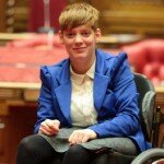 This is a picture of Ms Vincent in the South Australian Legislative Council. Ms Vincent has short red hair and is wearing a blue jacket. She is using a manual wheelchair and in front of the Clerk's benches and the Speaker's chair. Much of the Legislative Council decor around her is red, in accordance with the tradition in relation to upper houses in the Westminster system.