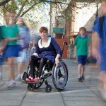 This is a picture of Ms Vincent in front of the South Australian Parliament. Ms Vincent has short red hair and is wearing a blue dress. She is sitting in a manual wheelchair. Children in blue and green school uniforms are walking past Ms Vincent, they are blurry due to the use of a long exposure effect.