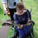 This is a photograph of Kelly Vincent holding a python at Mental Health Week. Ms Vincent is sitting in her manual wheelchair and is holding the snake in her hands just above her lap. The photograph has been taken above Ms Vincent so she is looking up at the camera and is smiling. She is wearing a blue floral print lace dress with dark pink lipstick. She is wearing spectacles with a black frame and has short red hair. In the background of the photograph is a person who is assisting with the snake. Green grass is also within the frame below and behind Ms Vincent.