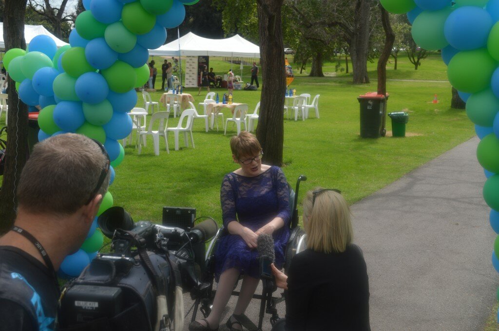 This is a photograph of Kelly Vincent being interviewed by ABC TV during Mental Health Week. Ms Vincent is sitting in her manual wheelchair in the centre of the photograph with a journalist sitting infront of her. A man holding a videocamera is also in frame on the left of the photograph. The interview is taking place in a park where there are balloons, chairs and stall set up in the background.