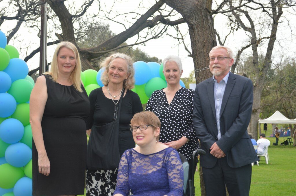 This is a photograph of Kelly Vincent at a Mental Health Week event with (from left to right) Hon Tammy Franks MLC, Dr Martha Kent, Ms Janne McMahon OAM, and Mr Geoff Harris. Ms Vincent is sitting at the front of the photograph in her manual wheelchair with everyone else standing behind her. They are are all facing the camera and are smiling. A park can be seen in the background as well as some balloons.