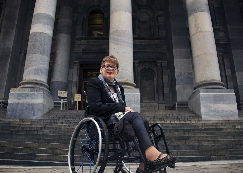 This is a photograph of Kelly Vincent in her manual wheelchair in front of the steps to Parliament House. She is in the centre of the photograph and is facing in the direction of the camera and smiling. She is wearing a white dress with a black bow and polka dot pattern. She has a black blazer on over the top of her dress and is also wearing black tights. She has a black and white gingham scarf around her neck and is wearing spectacles with a black frame. She is wearing dark pink red lipstick and has short red hair.