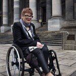 This is a photograph of Kelly Vincent in her manual wheelchair in front of Parliament House. She is facing in the direction of the camera and is smiling. She is wearing a white dress with a black bow and polka dot print. She is wearing a black blazer and black tights with a gingham black and white patterened scarf around her neck. She is also wearing spectacles with a black frame and has dark pink lipstick on. She has short red hair. Ms Vincent is sitting out the front of the steps at Parliament House.