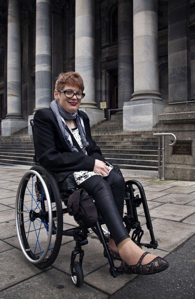 This is a photograph of Kelly Vincent in her manual wheelchair in front of Parliament House. She is facing in the direction of the camera and is smiling. She is wearing a white dress with a black bow and polka dot print. She is wearing a black blazer and black tights with a gingham black and white patterened scarf around her neck. She is also wearing spectacles with a black frame and has dark pink lipstick on. She has short red hair. Ms Vincent is sitting out the front of the steps at Parliament House.