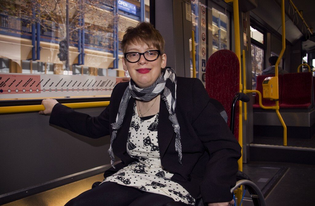 This is a photograph of Kelly Vincent riding the tram in her manual wheelchair. She is wearing a white dress with a black bow and polka dot pattern. She is also wearing a black blazer and black tights with a black and white gingham patterened scarf around her neck. She is wearing spectacles with a black frame and has dark pink lipstick on. She is holding on to the railing on the tram which is bright yellow.