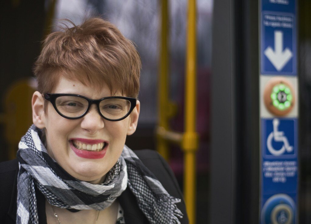 This is a photograph of Kelly Vincent alighting from a tram. She is looking in the direction of the camera and smiling broadly. Only Ms Vincent's head and shoulders are in the frame of the photograph. She is wearing black blazer and gingham patterened black and white scarf. She has spectacles on with a black frame and is also wearing deep pink lipstic. She has short red hair. In the background there is the door for the tram which has a button lit up on it for people to press and alight from the tram.