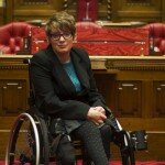 This is a photograph of Kelly Vincent sitting in her manual wheelchair in the Legislative Council Chamber, in front of the Clerk's desk. She is wearing a black blazer, blue crochet blouse, grey skirt and black stockings with a polka dot pattern. She is also wearing spectacles with a black frame and has red hair.