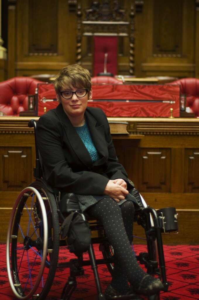 This is a photograph of Kelly Vincent sitting in her manual wheelchair in the Legislative Council Chamber, in front of the Clerk's desk. She is wearing a black blazer, blue crochet blouse, grey skirt and black stockings with a polka dot pattern. She is also wearing spectacles with a black frame and has red hair.