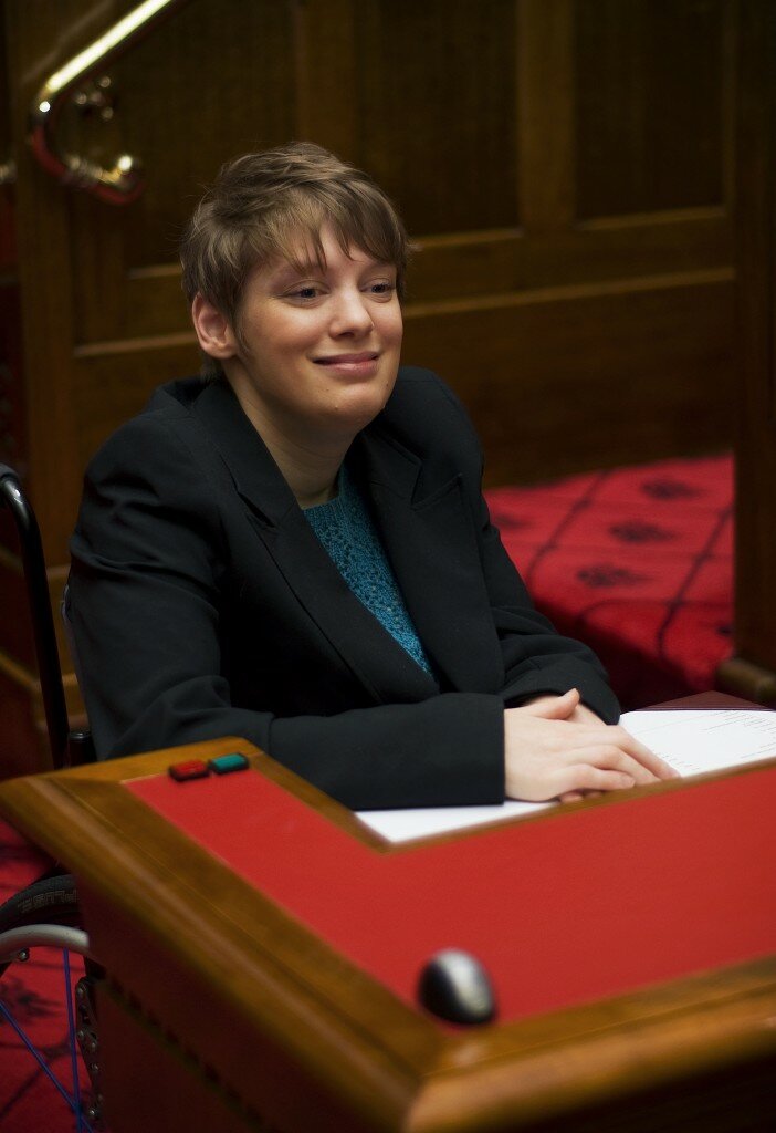 This is a photograph of Kelly Vincent sitting at her desk in the Legislative Council chamber. Ms Vincent is sitting in her manual wheelchair behind the desk with her hands resting on the desk. She is facing slightly to the right side of the camera and is smiling. She is wearing a black blazer with a blue crochet blouse. She has short red hair. Ms Vincent's wooden desk is at the front of the photograph with the red chamber carpet in the background behind Ms Vincent.