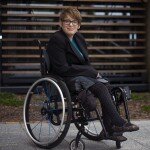 This is a photograph of Kelly Vincent sitting in the courtyard of Parliament House in her manual wheelchair. She is facing the camera and is smiling. Ms Vincent is wearing a black blazer with a blue crochet top, grey skirt and black stockings with a polka dot pattern. She is also wearing spectacles with a black frame and has short red hair. She is sitting in front of a small garden with some shrubs and bark chips, behind the garden is a large window.