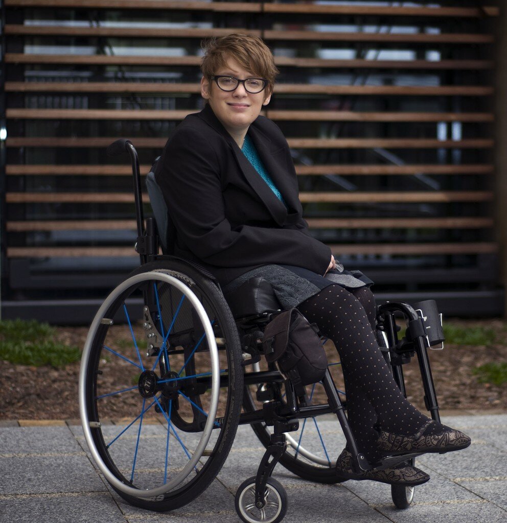 This is a photograph of Kelly Vincent sitting in the courtyard of Parliament House in her manual wheelchair. She is facing the camera and is smiling. Ms Vincent is wearing a black blazer with a blue crochet top, grey skirt and black stockings with a polka dot pattern. She is also wearing spectacles with a black frame and has short red hair. She is sitting in front of a small garden with some shrubs and bark chips, behind the garden is a large window.