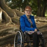 This is a photograph of Kelly Vincent sitting amongst the roots of a Morton Bay Fig Tree in Botanic Park in her manual wheelchair. She is in the centre of the photograph and is smiling in the direction of the camera. She is wearing a bright blue blazer with a yellow, pink, blue, black and white dress and black tights. She is wearing spectacles with a dark frame and has short red hair.