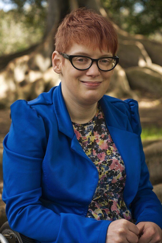 This is a photograph of Kelly Vincent in Botanic Park. She is sitting in her manual wheelchair and is looking in the direction of the photograph in smiling. Only the top half of Ms Vincent is in the frame of the photograph. She is wearing a bright blue blazer with a pink, yellow, blue, black and white floral dress. She is also wearing spectacles with a dark frame and has short red hair. Directly behind Ms Vincent is a large fig tree with it's very long roots above the ground.