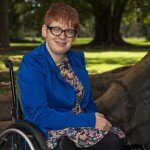 This is a photograph of Kelly Vincent in Botanic Park. Ms Vincent is facing the direction of the camera and smiling. She is sitting in her manual wheelchair with her hands in her lap. She is wearing a bright blue blazer with a yellow, pink, blue, black and white flora dress. She is wearing spectacles with a dark frame and has red hair. In the background is Botanic Park, there are multiple large trees behind Ms Vincent and green grass. Directly behind Ms Vincent are the very large roots of a tree she is sitting under.