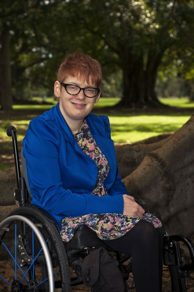 This is a photograph of Kelly Vincent in Botanic Park. Ms Vincent is facing the direction of the camera and smiling. She is sitting in her manual wheelchair with her hands in her lap. She is wearing a bright blue blazer with a yellow, pink, blue, black and white flora dress. She is wearing spectacles with a dark frame and has red hair. In the background is Botanic Park, there are multiple large trees behind Ms Vincent and green grass. Directly behind Ms Vincent are the very large roots of a tree she is sitting under.