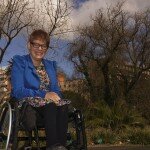 This is a photograph of Kelly Vincent in the Adelaide Botanic Gardens, with the Royal Adelaide Hospital visible in the background. Ms Vincent is facing the direction of the camera and smiling. She is sitting in her manual wheelchair with her hands in her lap. She is wearing a bright blue blazer with a yellow, pink, blue, black and white flora dress and black tights. She is wearing spectacles with a dark frame and has red hair. In the background there are multiple large trees behind Ms Vincent as well as some smaller shrubs and grass. The Royal Adelaide Hospital is also in the frame behind the trees. The sky is also within the frame of the photograph above the hospital which is a bright blue colour with some clouds.
