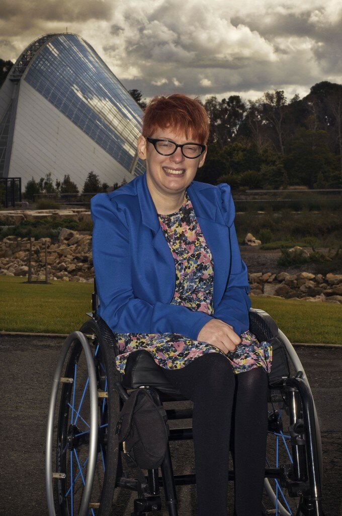This is a photograph of Kelly Vincent in her manual wheelchair in front of the Adelaide Botanic Garden's Tropical Conservatorium. Ms Vincent is in the centre of the photograph and is smiling in the direction of the camera. She is wearing a bright blue blazer with a yellow, pink, black, blue and white dress and black tights. She is also wearing spectacles with a dark frame and has short red hair.