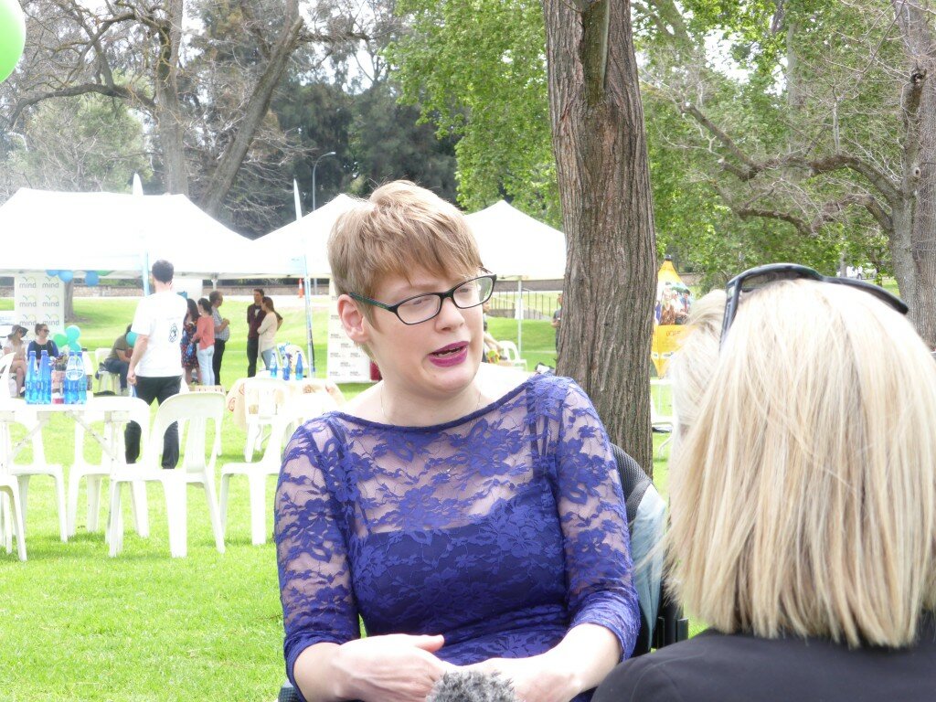 This is a photograph of Kelly Vincent speaking to the media at Mental Health Week. Ms Vincent is sitting in her manual wheelchair in the centre of the photograph. She is facing towards the camera but is looking in the direction of a news reporter who is sitting in front of Ms Vincent. Ms Vincent is wearing a blue floral patterend lace dress and dark pink lipstick. She is wearing spectacles with a black frame and has short red hair. The journalist Ms Vincent is speaking to in sitting on the right of the photograph. She is facing away from the camera and has short blonder hair. In the background of Ms Vincent is a park with white chairs and stalls set up. There are also some people gathered around in groups.