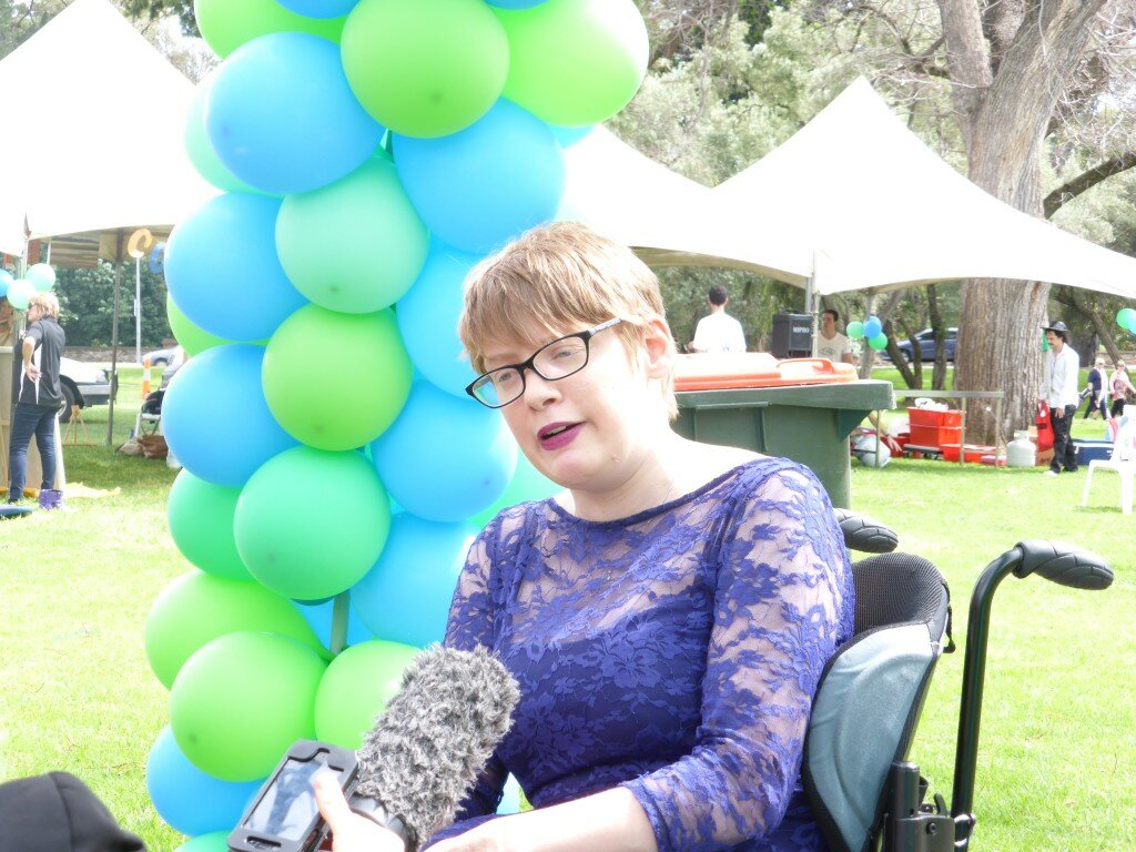 This is a photograph of Kelly Vincent speaking to media at Mental Health Week. She is sitting in her manual wheelchair in the centre of the photograph. She is facing to the left of the photograph towards the journalists interviewing her who are not in frame. She is wearing a blue floral lace dress and dark pink lipstick. She is also wearing spectacles with a black frame and has short red hair. In front of Ms Vincent a microphone and a mobile phone are being held out to record. Behind Ms Vincent is a park where there are balloons, and stalls set up.
