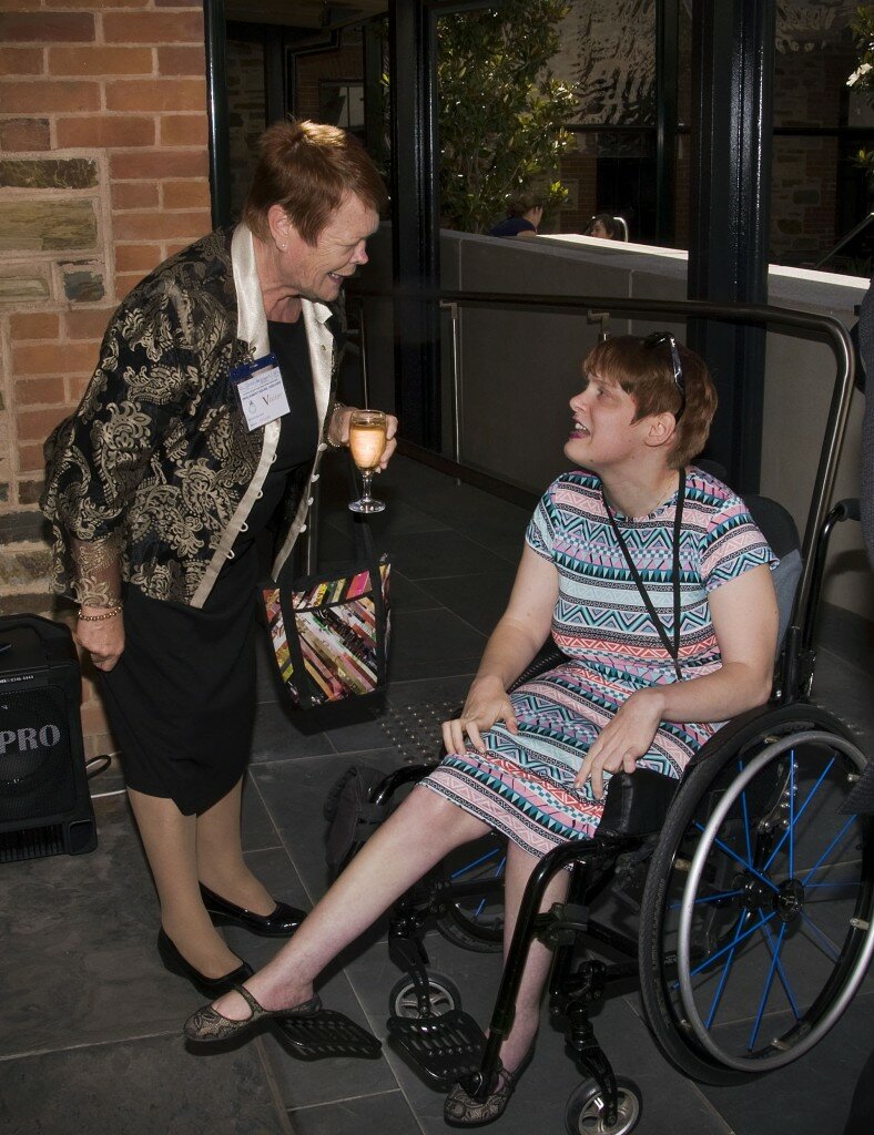 This is a photograph of Kelly Vincent with Glenys Jones OAM. Ms Jones is standing in the left of the photograph. She is leaning forward at the waist slightly and is looking at Ms Vincent and speaking. She is wearing a black dress, a black jacket with a large golden paisley print and light grey collar and buttons, and shiny black leather shoes. Around her right wrist is a gold bracelet and is carrying a handbag with bright multi-coloured diagonal line pattern at her left elbow and a wine glass full of white wine in her left hand. She has short red-brown hair and is wearing purple lipstick. Ms Vincent is in the right of the photograph and is seated in her manual wheelchair, which has a black frame and light blue spokes on it's wheels. She is wearing a multi-coloured dress with a bold print of triangles in reds, greens, pinks, and black and white. Around her neck hangs a black lanyard with an id hanging from it and her black framed Wayfarer sunglasses have been pushed back onto the top of her head. She has short red hair and is wearing dark mauve lipstick. She is looking at Ms Jones and speaking with her. Her hands rest on her knees. In the background of the photograph is the blue stone wall of Old Parliament House and a accessibility ramp with a metal handrail and tactile pathway indicators. further away, in the courtyard, small potted trees are visible.
