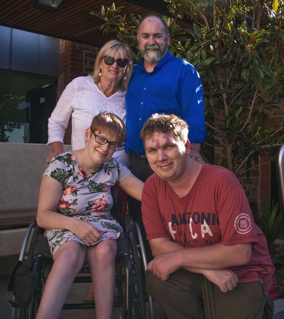 This is a photography of Colleen Hunt, Geoffrey Hale, Kelly Vincent and Shane Vincent. Mrs Hunt and Mr Hale are standing at the back of the photograph. They are both looking in the direction of the photograph and smiling. Ms Hunt is wearing dark sunglasses and white embroidered blouse. She has shoulder length blonde hair. Mr Hale is wearing an electric blue button up shirt. He has short dark grey hair and a beard. Ms Vincent and Mr Vincent are sitting at the front of the photograph. They are both looking in the direction of the camera and smiling. Ms Vincent is sitting in her manual wheelchair.She is wearing a light blue dress with a pink, white, and green floral print featuring hibiscus and orchids. She is wearing spectacles with dark frames and has short, red hair. Mr Vincent is squatting down on the right of Ms Vincent. He is wearing a red t-shirt with dark green pants. He has short brown hair. In the background of the photograph on the right hand side behind Mr Hale is a small tree. On the left hand side, the building can be seen which is made up on red bricks and grey walls.