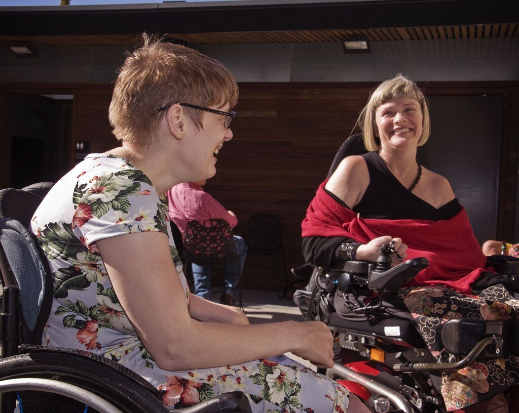 This is a photograph of Kelly Vincent and Joanne Blesing. Ms Vincent is seated to the left of the photograph in her manual wheelchair. She is sitting side on and is quite close to the camera. She is wearing a light blue dress with a pink, white, and green floral print featuring hibiscus and orchids. She is wearing spectacles with dark frames and has short, red hair. She is looking away from the camera and to the right, in the direction of Joanne Blesing, and is laughing. Ms Blesing is seated to the right of the photograph and is slightly further away from the camera. She is sitting in a large, red, powered wheelchair. She is wearing a dark navy dress with a bright, geometric print, based on a Japanese floral crest, and a red pashmina. She has blonde hair styled into a bob, and is smiling broadly. The in the background of the photograph is a timber wall with a verandah projecting out from it. Two grey doors in the wall are visible in the far left and right of the picture, and the door to the left is slightly ajar, opening inwards. A man in a pink shirt and blue jeans can be seen sitting in a black plastic designer outdoor chair near the middle of the photograph, his face obstructed by Ms Vincent's.