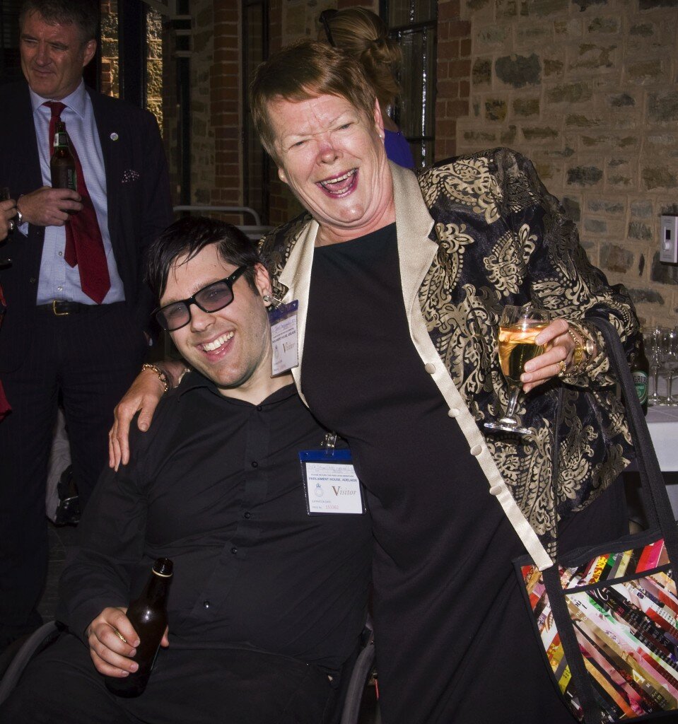 This is a photograph of Nick Schumi and Glenys Jones OAM. Mr Schumi is seated in his manual wheelchair to the left of the photograph. He is wearing a black business shirt and black trousers and is holding a bottle of ginger ale in his right hand. He has medium length black hair parted on the left and swept across, and is wearing spectacles with thick black frames and lenses with a slight purple tint. He is smiling broadly. Ms Jones is standing to the left of the picture. She is weating a black dress and a black jacket with a large, golden paisley pattern and a light grey collar. She has her a handbag with a bright multi-coloured diagonal line design hooked on her left elbow and is holding a full glass of white wine in her left hand. She has short, light brown hair that is styled to the left. She is leaning with her right arm around Mr Schumi's shoulders and is laughing uproariously. In the background of the photograph the bluestone wall of Old Parliament House is visible, in front of which is a table laden wine glasses and bottles of soft drink. To the left of the picture Dignity for Disability President Rick Neagle is in conversation, wearing a navy pinstripe suit, light blue shirt and red tie. He is holding a bottle of beer in his right hand and has short, light brown hair which is greying slightly. He is looking to the left, in the direction of someone who is out of frame.