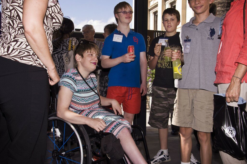 This is a photograph of Kelly Vincent with Ben Paior-Smith, Bailey Paior-Smith and one of their friends. Ms Vincent is sitting in her manual wheelchair in the centre of the photograph. She is wearing a bright, multi-coloured dress featuring a bold triangle pattern in greens, pinks, reds, and black and white. She has a black land yard around her neck and dark sunglasses siting on top of her head. She is also wearing bright pink lipstick and has short red hair. She is facing the camera but is looking away and smiling. On the right of Ms Vincent is Ben Paior-Smith. He is wearing a dark blue polo shirt with red shorts. He is wearing spectacles with light purple lenses and has short blonde hair. He is holding a can of soft drink and is also facing in the direction of the camera but looking away and smiling. On the right of the Ben Paior-Smith is one of his friends. His friend is wearing a black t-shirt and camouflage print shorts. He is also holding a soft drink. He is facing the camera but is look down. On the right hand side is Bailey Paior-Smith. He has been cut out of the photograph so only half of his face is visible. He is wearing a grey polo shirt and light brown shorts. Sam Paior is also visible in this photograph on the left hand side, however her face has been cut out. She is wearing a white and brown top with a floral and leaf patter and black pants.