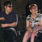 This is a photograph of Nick Schumi and Kelly Vincent. Mr Schumi is sitting to the left of the photograph in his manual wheelchair. He is wearing a dark purple collared shirt with black trousers. He is wearing spectacles with a dark frame and purple lenses. He has short black hair. He is using both hands to open a beer that is resting on his lap. Ms Vincent is sitting to the right of the photograph in her manual wheelchair. She is wearing a light blue dress with a pink, white, and green floral print featuring hibiscus and orchids. She is wearing spectacles with dark frames and has short, red hair. They are both looking away from the camera in the direction of something out of frame on their left.