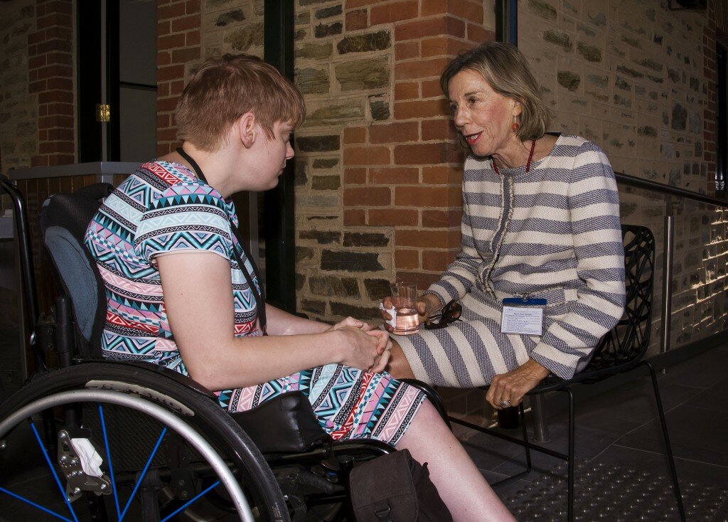 This is a photograph of Kelly Vincent and Lesley Brydon. Ms Vincent is sitting in her manual wheelchair on the left hand side of the photograph. She is wearing a bright, multi-coloured dress featuring a bold triangle pattern in greens, pinks, reds, and black and white. She has a black land yard around her neck and has short red hair. She is facing away from the camera in the direction of Ms Brydon. Ms Brydon is sitting in a black chair on the right hand side of the photograph. She is wearing a navy and cream striped dress. She is also wearing a red beaded necklace and earings. She is holding a glass of water in her right hand and resting it on her lap. She has blonde hair which sits just above her shoulders. She is facing towards Ms Vincent and is speaking. In the background of the photograph is a wall which is made up of assorted bricks.