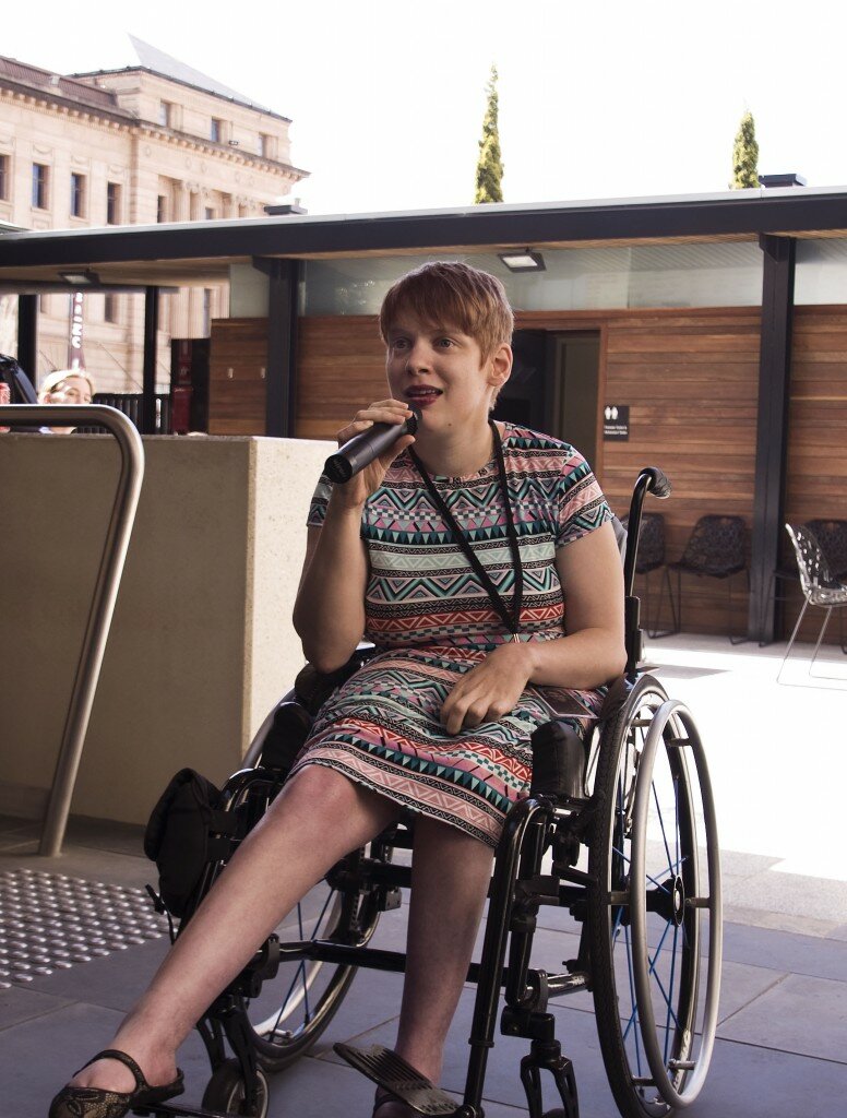 This is a photograph of Kelly Vincent. Ms Vincent is seated in her manual wheelchair in the courtyard of Parliament House. She is wearing a bright, multi-coloured dress featuring a bold triangle pattern in greens, pinks, reds, and black and white. She has a black lanyard around her neck and is speaking into a microphone which she holds in her right hand. Her left hand rests in her lap. She is looking past the camera, up and slightly to the left. In the background of the picture is the imposing sandstone wall of the Adelaide Railway Station. Nearer the camera there is a wooden wall from which projects a small verandah supported by thigh, black painted, metal beams. A number of clack, plastic, designer outdoor chairs sit on the grey pavers behind Ms Vincent. To the left of the picture is a short, off-white, cement diving wall, which roughly lines up with the top of Ms Vincent's right shoulder. In front of the wall a brushed steal handrail rises from the ground, turns towards the left and continues out of frame.
