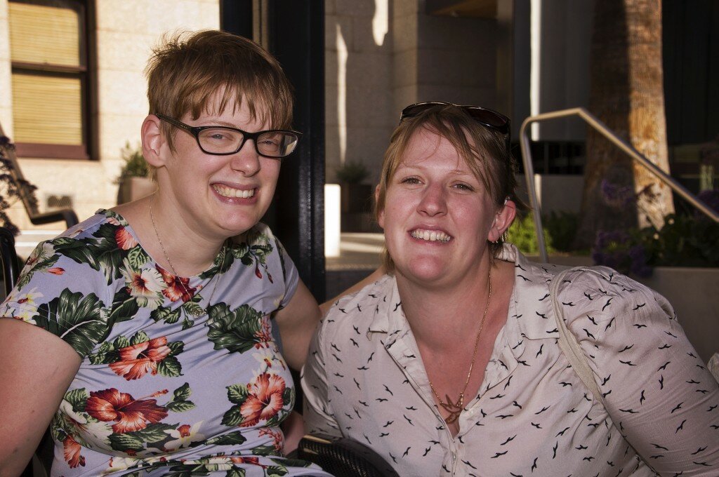This is a photograph of Kelly Vincent and Bonnie Millen. Ms Vincent is sitting in her manual wheelchair on the left hand side of the photograph. She is wearing a light blue dress with a pink, white, and green floral print featuring hibiscus and orchids. She is wearing spectacles with a dark frame and has short red hair. She is smiling broadly and is looking in the direction of the camera with her arm around Ms Millen. Ms Millen is on the right of the photograph. She is wearing white blouse that has a small black sparrow pattern on it. She has a pair of dark glasses on her head and is also wearing a gold sparrow necklace. She has short light brown hair. She is also smiling broadly and looking in the direction of the camera.