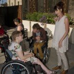This is a photograph of Kelly Vincent, Laurence Byrne, and Ingrid Birgden. Ms Vincent is sitting in her manual wheelchair on the left of the photograph. She is wearing a light blue dress with a pink, white, and green floral print featuring hibiscus and orchids. She is wearing spectacles with dark frames and has short, red hair. She is looking away from the camera towards Ms Birgden. Ms Birgden is standing on the right of the photograph. She is wearing a long cream singlet with cream trousers and brown boots. She has long brown hair that has been tied up into a bun. She is smiling broadly and is looking at Ms Vincent. Mr Byrne is sitting in his electric wheelchair in the middle of the photograph. He is wearing a black and white striped shirt with a red hooded jumper and mustard yellow jeans. He has short blonde hair and sitting with his hands in his lap. He is looking away from the camera to his righthand side.
