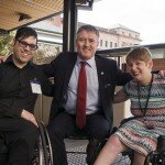This is a photograph of Nick Schumi, Rick Neagle, and Kelly Vincent. Mr Schumi and Ms Vincent are seated in their manual wheelchairs, and Mr Neagle sits between them on a black plastic designer outdoor chair. Mr Neagle has his hands around the shoulders of Mr Schumi and Ms Vincent and they each have an arm around Mr Neagle's back. All three are looking into the camera and smiling broadly. Mr Schumi is to the right of the photograph and is wearing a black business shirt and black trousers. His black, medium-length hair is swept across to his right and he is wearing spectacles with thick black lenses and lenses with a subtle purple tint. His right hand rests on his left knee. Mr Neagle is in the centre of the photograph. He is wearing a navy pinstripe suit with a light blue shirt and a red tie. he Has a small badge on his left lapel and a navy handkerchief with a polka dot pattern in his left breast pocket. He has short light brown, greying hair. Ms Vincent is to the right of the photograph and is wearing a brightly coloured dress with a bold triangle patterns in pinks, greens, reds, and black and white. An id card on a black lanyard hangs around her neck and her left hand rests on the grip of her wheelchair's left wheel. She has short red hair and is wearing dark pink lipstick.