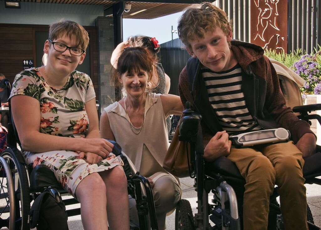 This is a photograph of Kelly Vincent, Ingrid Birgden and Laurence Byrne. They are all looking in the direction of the camera and smiling. Ms Vincent is sitting on the left of the photograph in her manual wheelchair. She is wearing a light blue dress with a pink, white, and green floral print featuring hibiscus and orchids. She is wearing spectacles with dark frames and has short, red hair. Ms Birgden is squatting down between Ms Vincent and Mr Byrne. She is wearing a cream, collared top with cream pants. She has dark brown hair with a fringe and has clipped her hair up. Mr Byrne is on the right of the photograph and is sitting in his electric wheelchair. He is wearing a black and white striped t-shirt with a red hooded jumper and mustard yellow pants. He has short blonde hair. In the background of the photograph on the right hand side is a timber fence with a small garden in front. On the left hand side in the background is a building made up of grey bricks and dark timber panels.