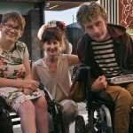 This is a photograph of Kelly Vincent, Ingrid Birgden and Laurence Byrne. They are all looking in the direction of the camera and smiling. Ms Vincent is sitting on the left of the photograph in her manual wheelchair. She is wearing a light blue dress with a pink, white, and green floral print featuring hibiscus and orchids. She is wearing spectacles with dark frames and has short, red hair. Ms Birgden is squatting down between Ms Vincent and Mr Byrne. She is wearing a cream, collared top with cream pants. She has dark brown hair with a fringe and has clipped her hair up. Mr Byrne is on the right of the photograph and is sitting in his electric wheelchair. He is wearing a black and white striped t-shirt with a red hooded jumper and mustard yellow pants. He has short blonde hair. In the background of the photograph on the right hand side is a timber fence with a small garden in front. On the left hand side in the background is a building made up of grey bricks and dark timber panels.