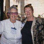 This is a photograph of Sandra Dann and Glenys Jones OAM, both of whom are looking into the camera and smiling. Ms Dann is standing to the left of the photograph and is wearing a white shirt with a decorative yoke. She is wearing spectacles with thick purple rims and a thin black necklace. Her short, silver hair is spelt up into a peak. Ms Jones stands to the right of the photograph and is wearing a black shirt and a silky black jacket with a large, gold, paisley pattern and light grey collar and buttons. She has short red-brown hair swept to her left and is wearing dark pink lipstick and small pearl earrings. In the background the light grey granite wall of Parliament House is visible, in front of which are raised beds of purple flowers and other guests gathered to speak with one another.