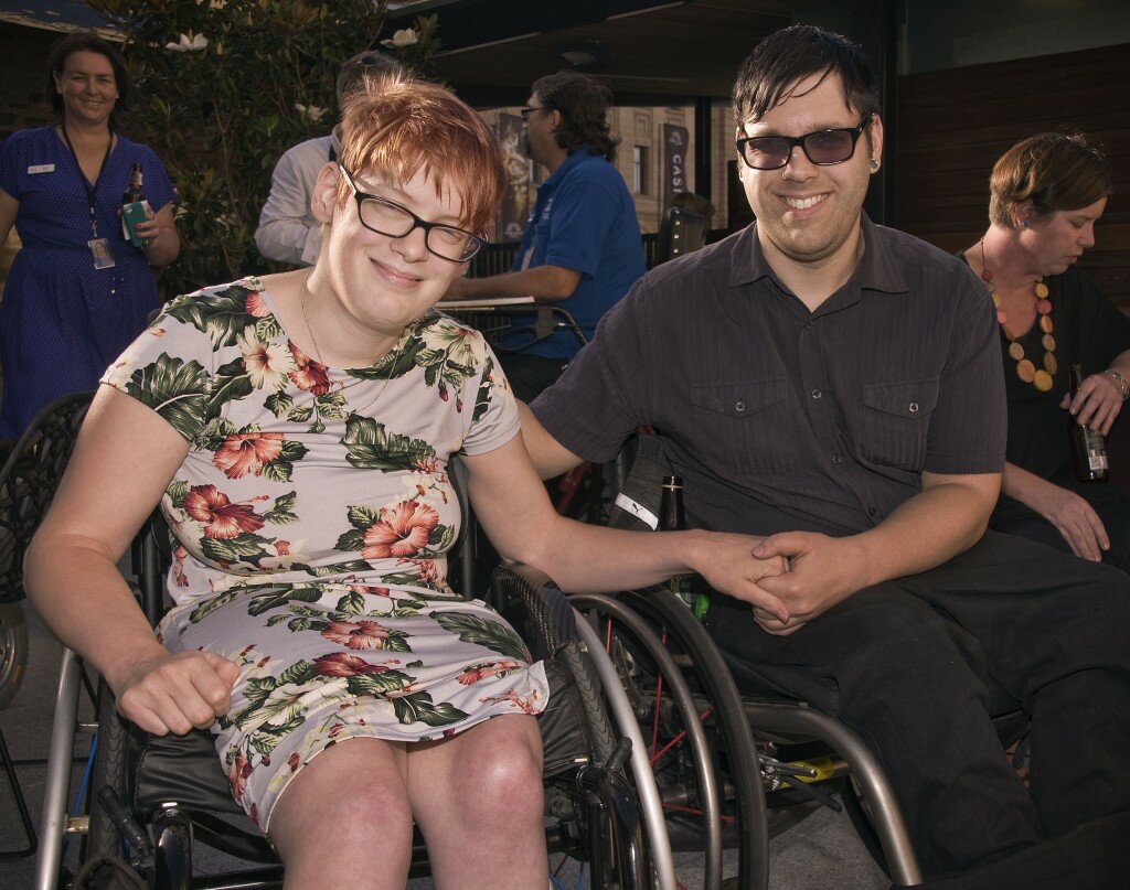 This is a photograph of Kelly Vincent and Nick Schumi. Ms Vincent is sitting in her manual wheelchair on the left hand side of the photograph. She is wearing a light blue dress with a pink, white, and green floral print featuring hibiscus and orchids. She is wearing spectacles with a dark frame and has short red hair. She is smiling and is looking in the direction of the camera. Mr Schumi is sitting in his manual wheelchair on the right side of the photograph. He is wearing a dark purple collared shirt and black trousers. He is wearing spectacles with dark purple lenses. He has short black. He is smiling broadly and looking in the direction of the camera. Mr Schumi and Ms Vincent are holding hands.