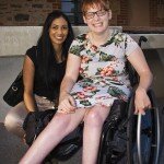 This is a photograph of Sana Ali and Kelly Vincent. Sana Ali is sitting on the left of the photograph. She is wearing a black t-shirt with bronze satin pants. She has long black hair. She is smiling broadly and looking in the direction of the camera. Ms Vincent is sitting in her manual wheelchair on the right of hte photograph. She is wearing a light blue dress with a pink, white, and green floral print featuring hibiscus and orchids. She has short red hair. She is smiling broadly and is looking in the direction of the camera.