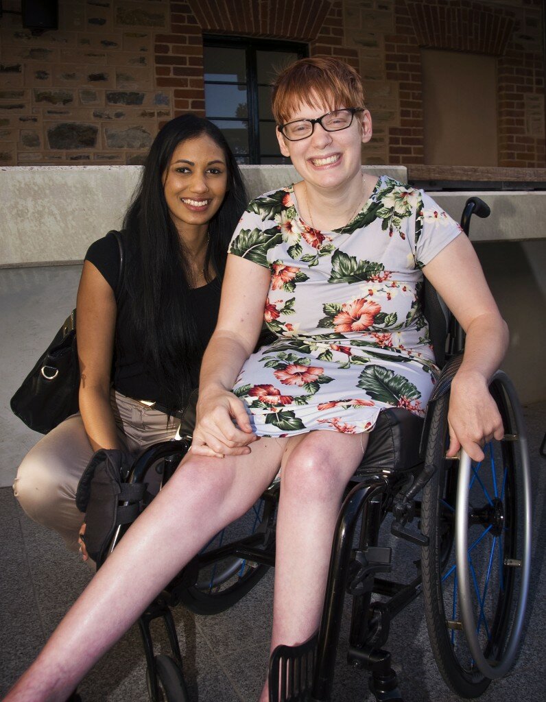 This is a photograph of Sana Ali and Kelly Vincent. Sana Ali is sitting on the left of the photograph. She is wearing a black t-shirt with bronze satin pants. She has long black hair. She is smiling broadly and looking in the direction of the camera. Ms Vincent is sitting in her manual wheelchair on the right of hte photograph. She is wearing a light blue dress with a pink, white, and green floral print featuring hibiscus and orchids. She has short red hair. She is smiling broadly and is looking in the direction of the camera.