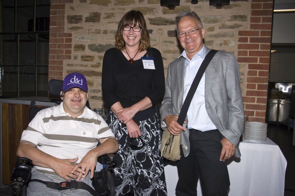 This is a photograph of Phillip Beddall, Anne Bainbridge, and Haydon Manning. They are standing in a row in front of the blue stone wall of Old Parliament House. THey are all looking into the camera and smiling. Mr Beddall is in the left of the photograph, seated in his black powered wheelchair. He is wearing a white polo shirt with a small embroidered penguin on the left breast and is patterned with narrow, horizontal tan lines that are widely spaced and light grey trousers. On his head is a purple baseball cap with a white embroidered logo which reads "d4d" in large type, with the words "Dignity for Disability" beneath it in smaller type. A seatbelt rests loosely around his hips and his hands rest folded in his lap. Next to Mr Beddall, in the centre of the photograph, stands Anne Bainbridge. Ms Bainbridge is wearing a black shirt, a black cardigan with 3/4 length sleeves, and a black skirt with a white marbled pattern. around her neck is a thick black necklace decorated with a large red-brown pendant. She is wearing spectacles with thick, black rims and has shoulder length, light brown hair, which is lighter at the ends. Her arms rest in front of her at her waist, her left hand holding her right wrist, her right wrist holding a pair of sunglasses. To the right of the photograph, next to Ms Bainbridge, is Haydon Manning. He is wearing very pale blue business shirt, light grey suit jacket, and black trousers. He is wearing spectacles with thin, light coloured metal frames and has short, silver hair. Around his body he wears a satchel, with the thick black strap over his left shoulder and the stone coloured bag resting on his right hip. His right hand gently grips the strap near where it joins the body of the bag, his left hand resting at his side with his thumb hooked in the pocket of his trousers.Behind them is a table with a white table cloth, which is laden with drinking glasses and small, white, china plates.