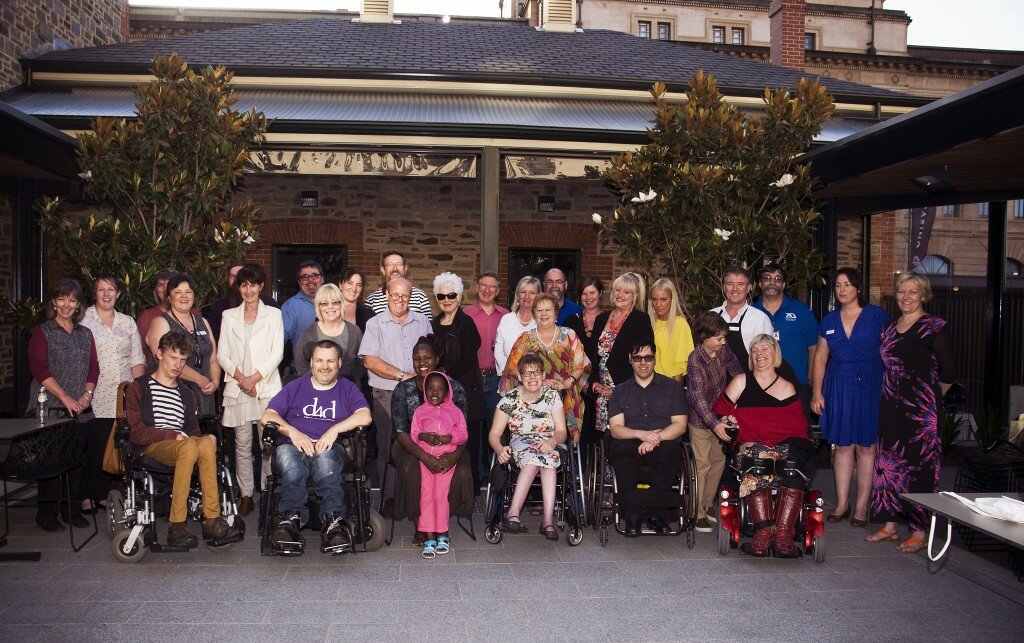 Members of Dignity for Disability gathered for end of year celebrations. There are 30 people pictured in this photograph who are all facing in the direction of the camera for the photograph. The group are gathered in the terrace with a brick building in the background