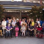 Members of Dignity for Disability gathered for end of year celebrations. There are 30 people pictured in this photograph who are all facing in the direction of the camera for the photograph. The group are gathered in the terrace with a brick building in the background